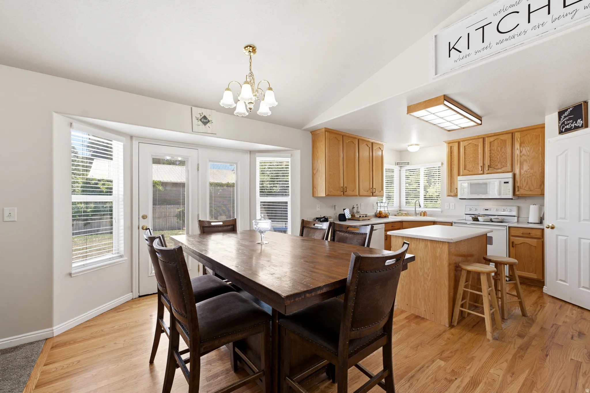 Open-concept kitchen and dining area featuring wood-finish flooring, light wood cabinetry, a white countertop island, and a bay window with a glass-paneled door