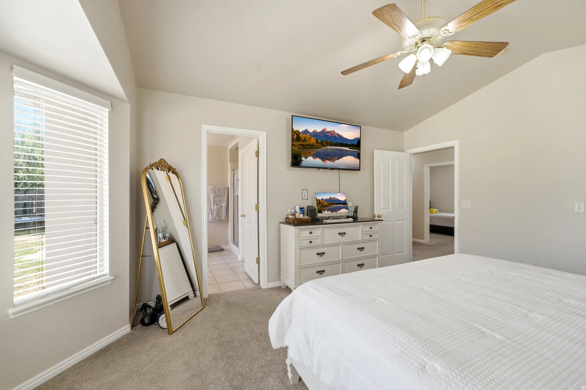 Carpeted bedroom featuring a vaulted ceiling with a ceiling fan, a window with horizontal blinds, and a doorway opening to a tiled bathroom