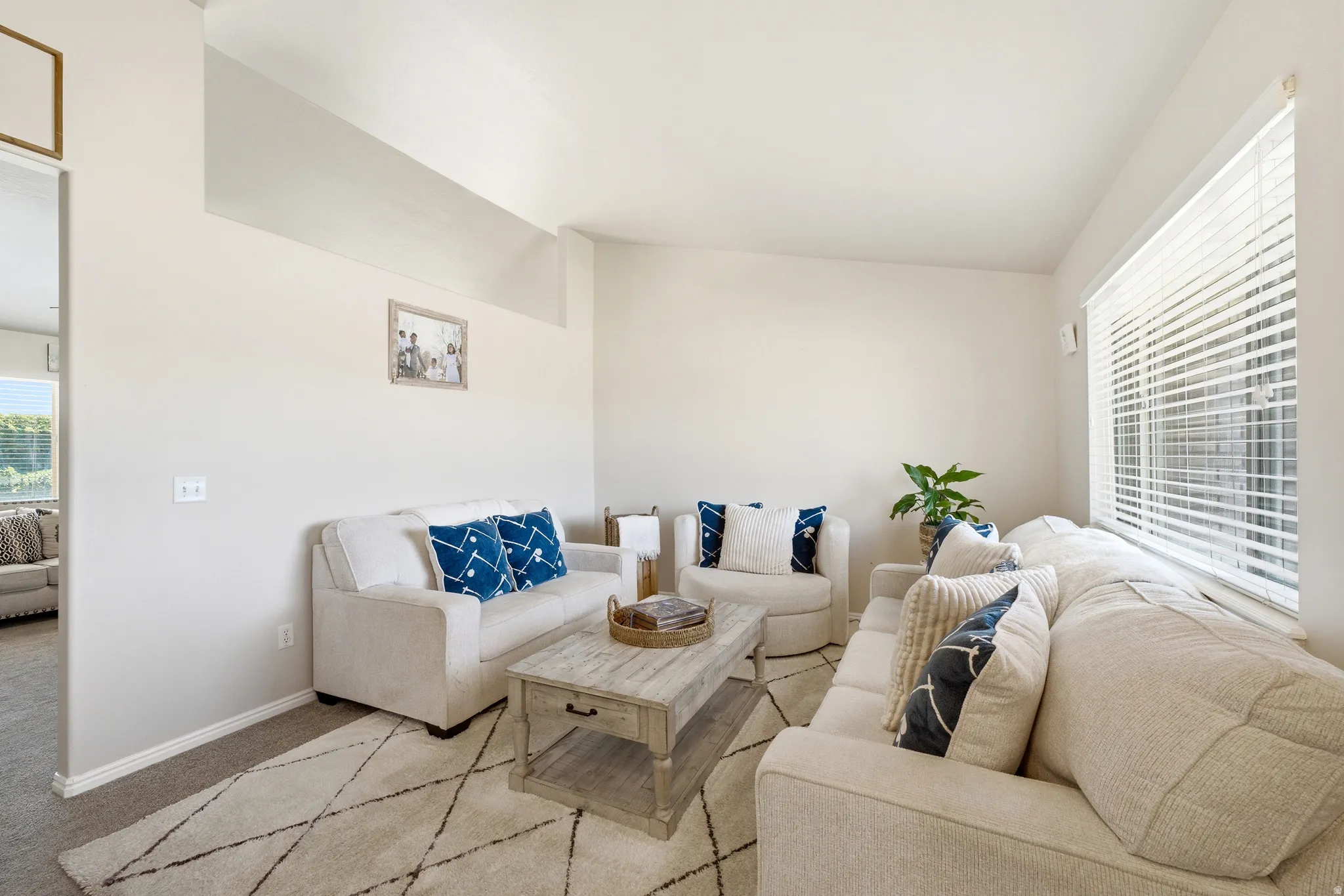 Spacious living area featuring light-toned walls, a large window with horizontal blinds, and carpeted flooring