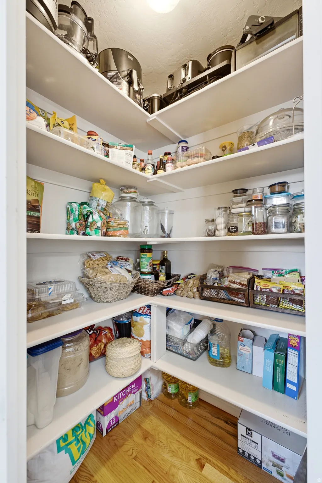 Pantry featuring white built-in shelving, corner storage, wood-finish flooring, and overhead lighting