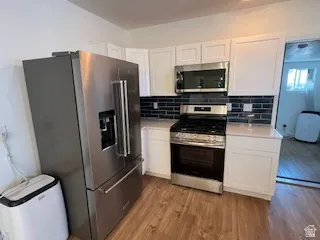 Modern kitchen featuring stainless steel appliances, white cabinetry, a dark subway tile backsplash, and light wood-finish flooring