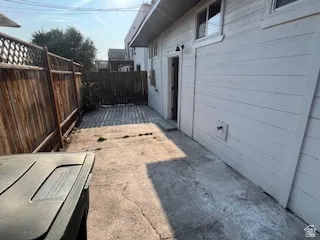 Paved side yard featuring a wooden privacy fence, horizontal siding, and a doorway with an exterior light fixture
