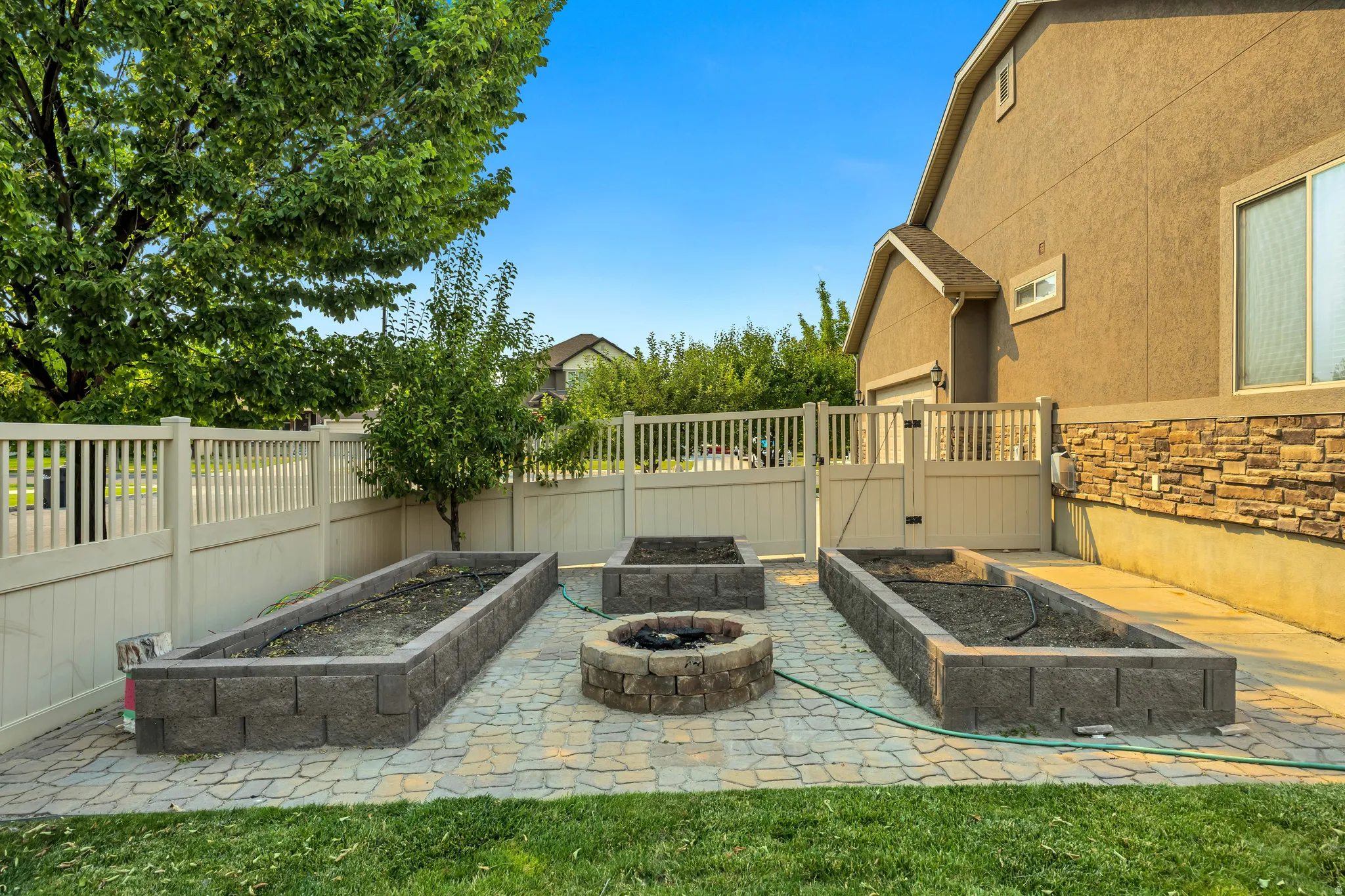 Paved patio featuring a fire pit, three raised garden beds, and a perimeter vinyl fence