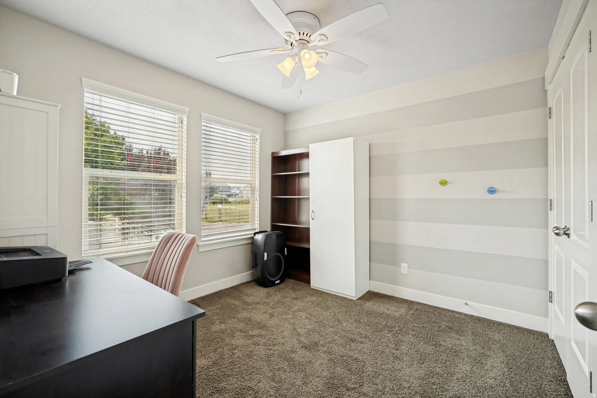 Carpeted room featuring two windows with blinds, a ceiling fan with integrated lighting, and a neutral striped accent wall