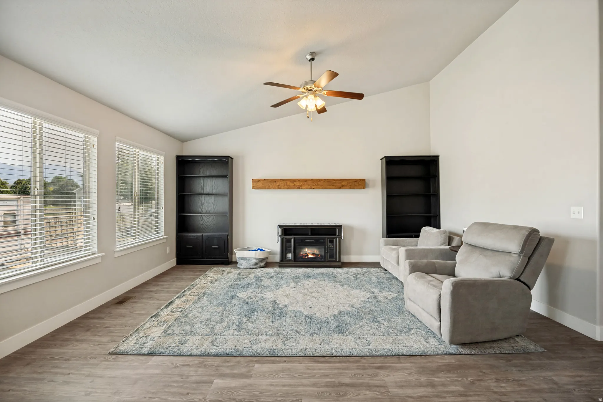 Vaulted ceiling living area featuring wood-finish flooring, multiple windows with blinds, a ceiling fan, and a built-in electric fireplace with a wood mantle