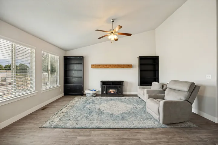 Vaulted ceiling living area featuring wood-finish flooring, multiple windows with blinds, a ceiling fan, and a built-in electric fireplace with a wood mantle