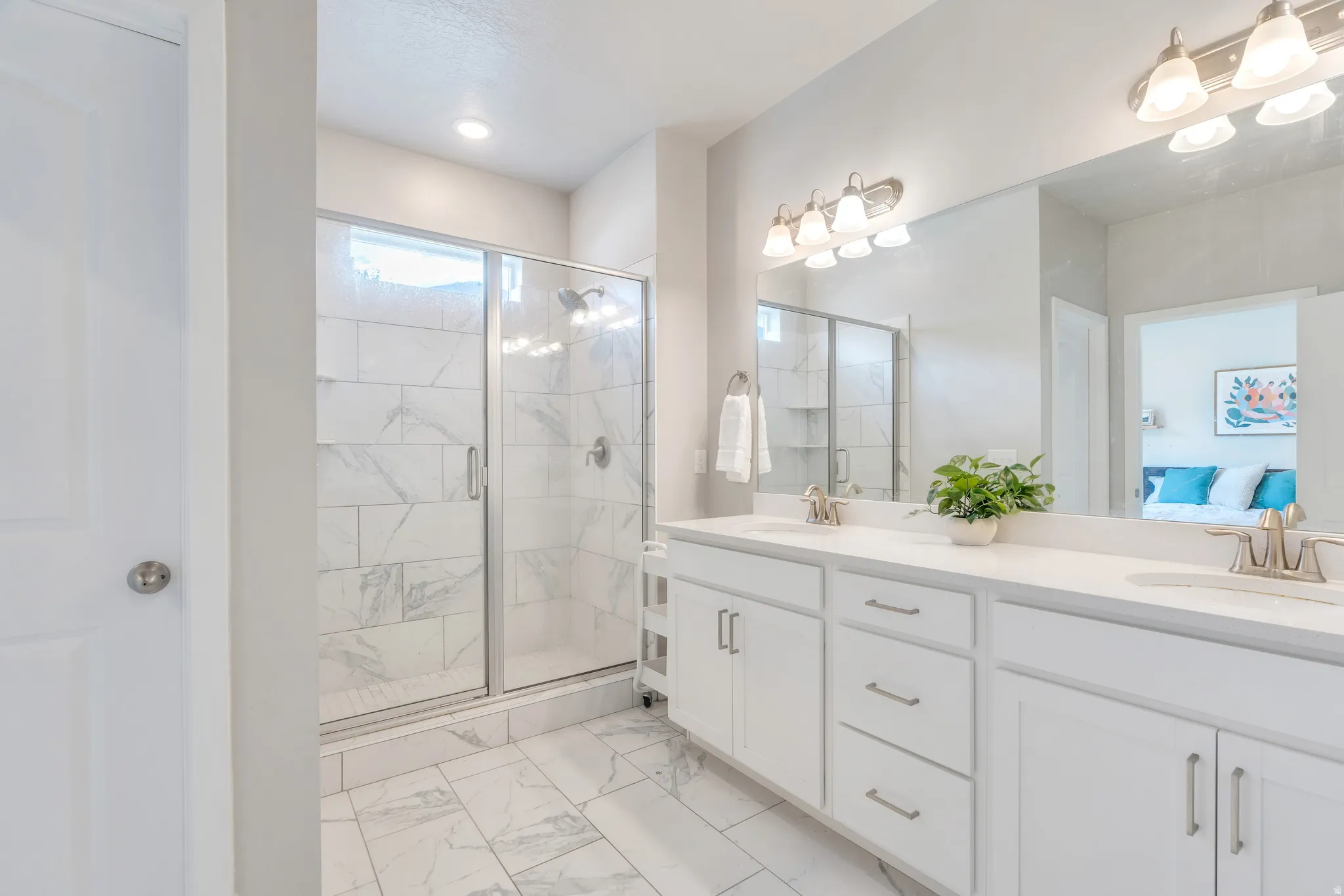 Spacious bathroom featuring a double vanity with stone-finish countertops, white cabinetry, a large frameless mirror, and a glass-enclosed shower with tiled walls and flooring