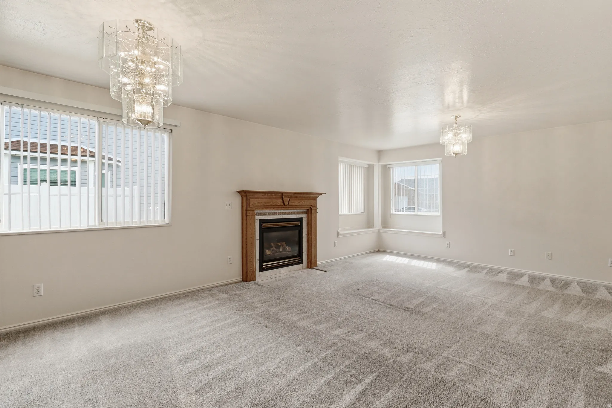 Spacious room featuring a central fireplace with a wood-finish mantel, light gray carpeting, and multiple windows with vertical blinds