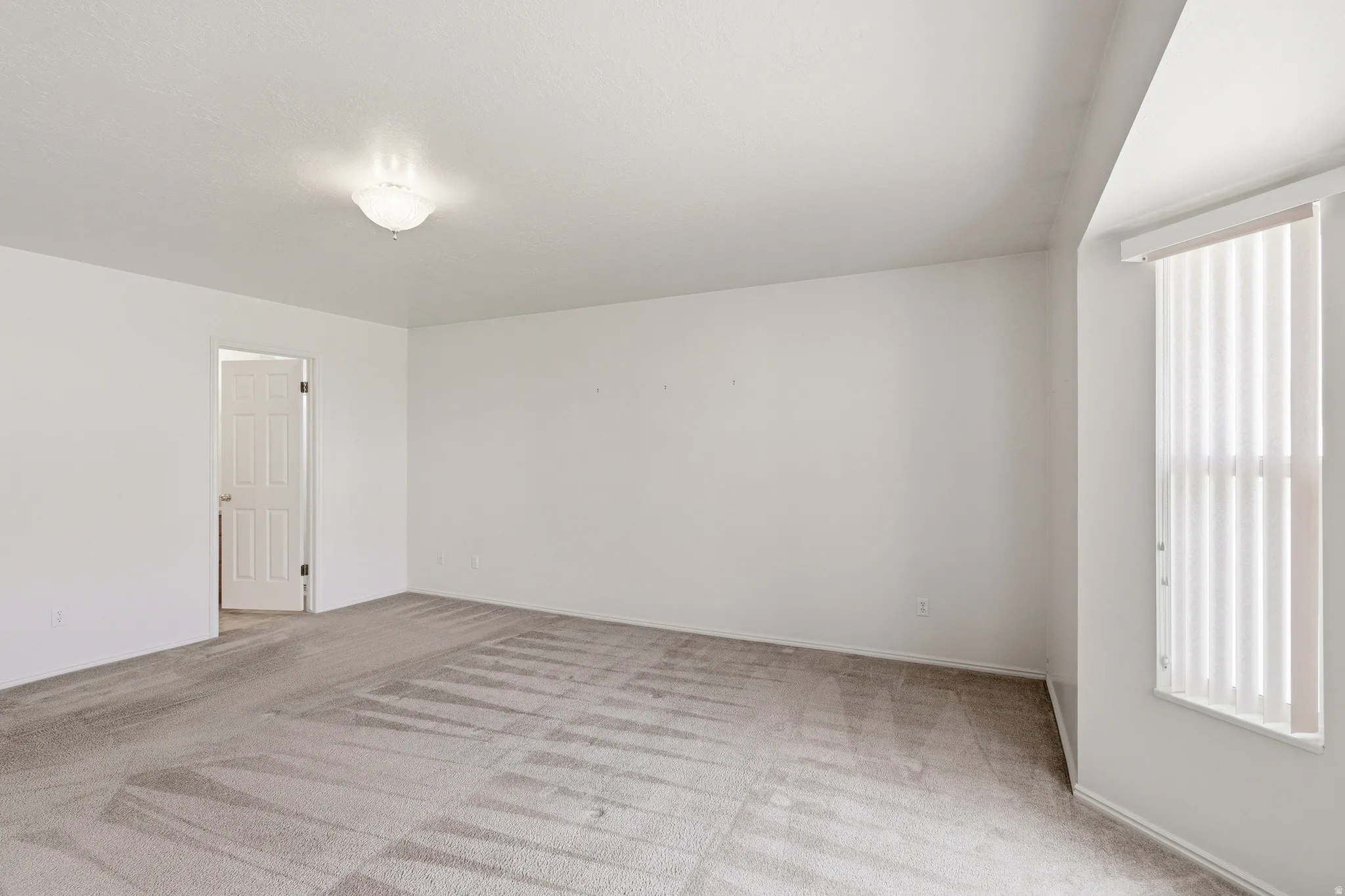 Spacious room featuring neutral carpeting, white walls, a flush-mount ceiling light, and a bay window with vertical blinds
