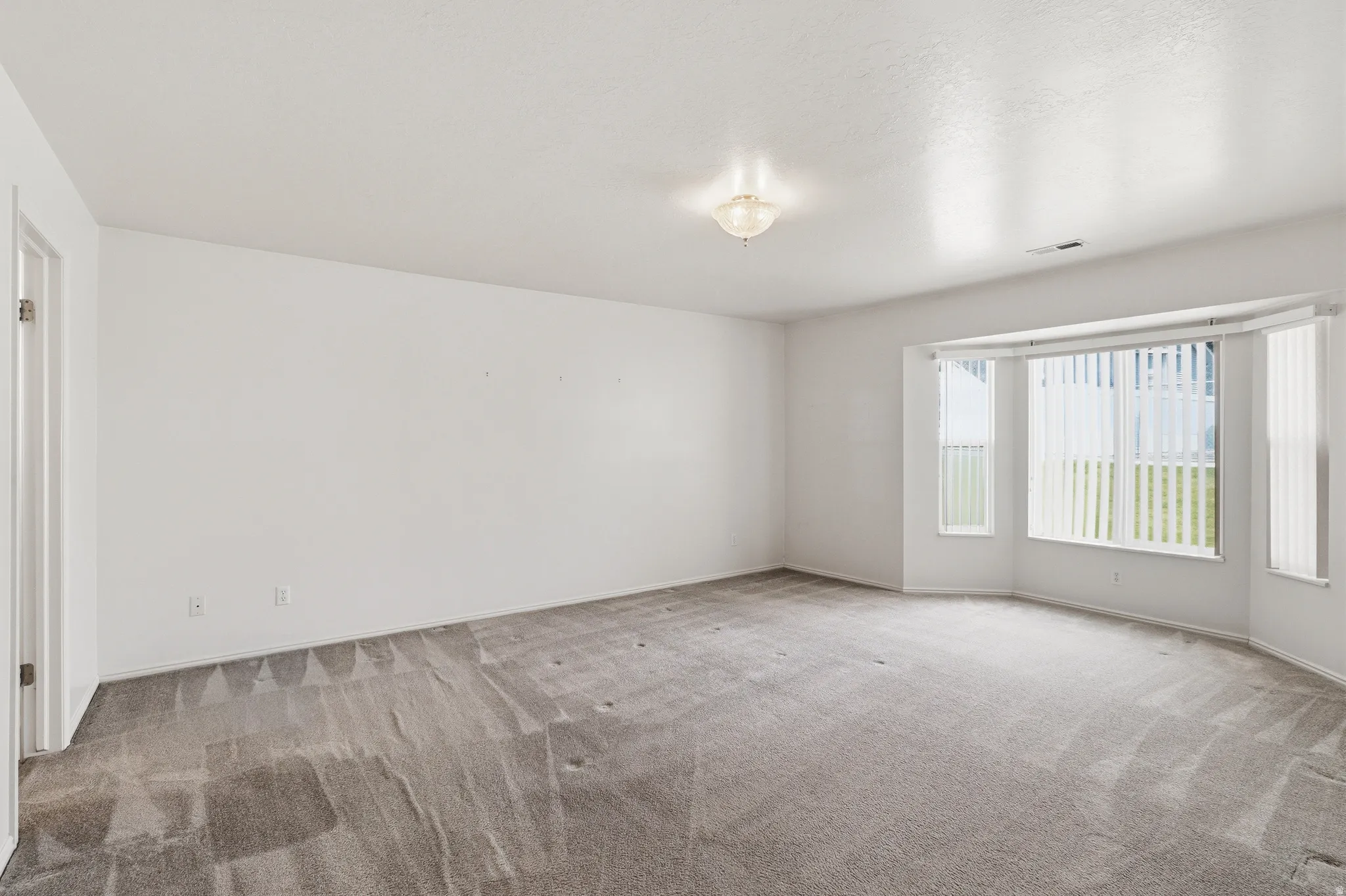 Spacious room featuring a bay window with vertical blinds, light gray carpet, and white walls