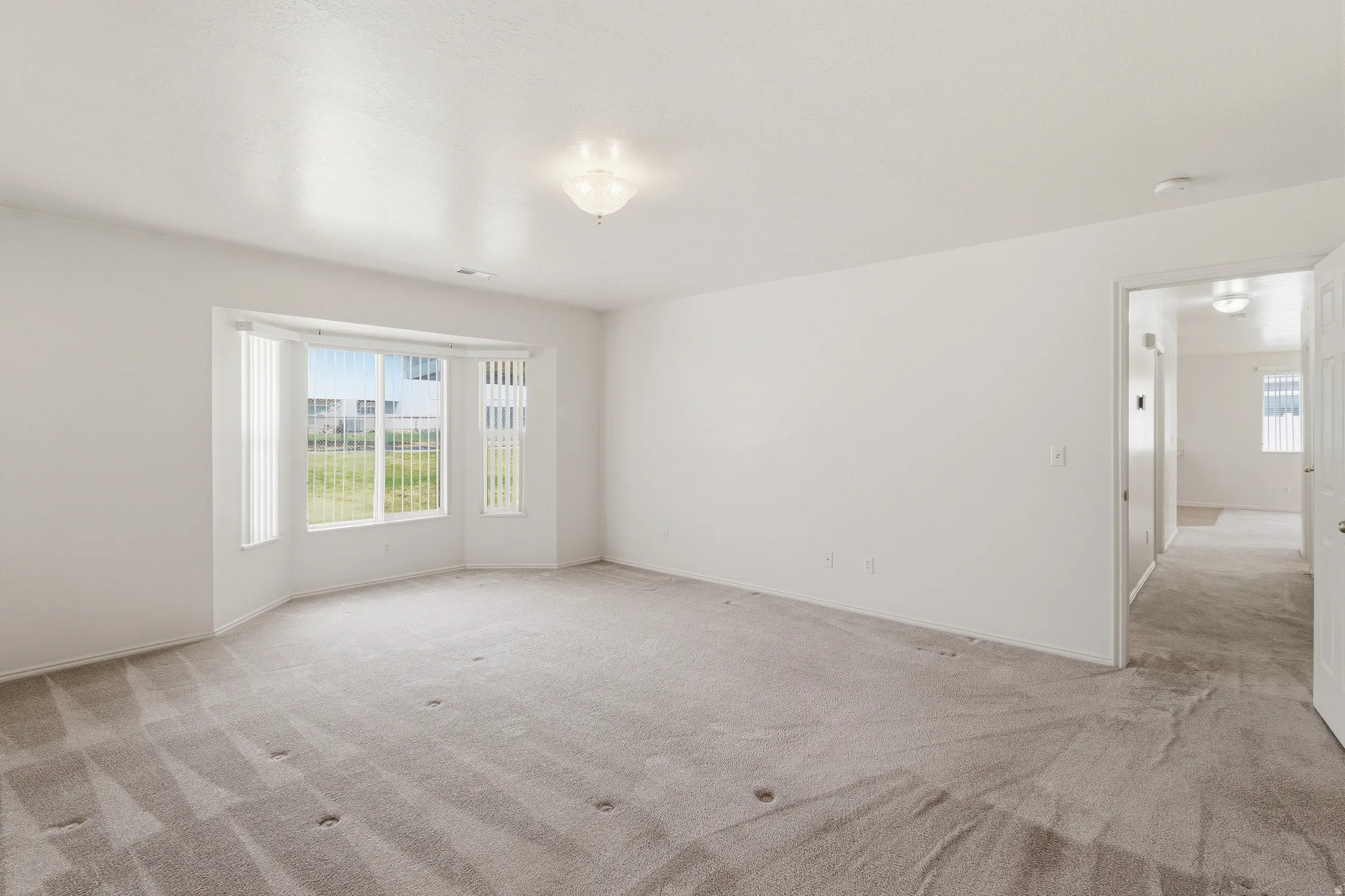 Spacious room featuring a bay window with vertical blinds, light grey carpeting, white painted walls, and a semi-flush mount ceiling light