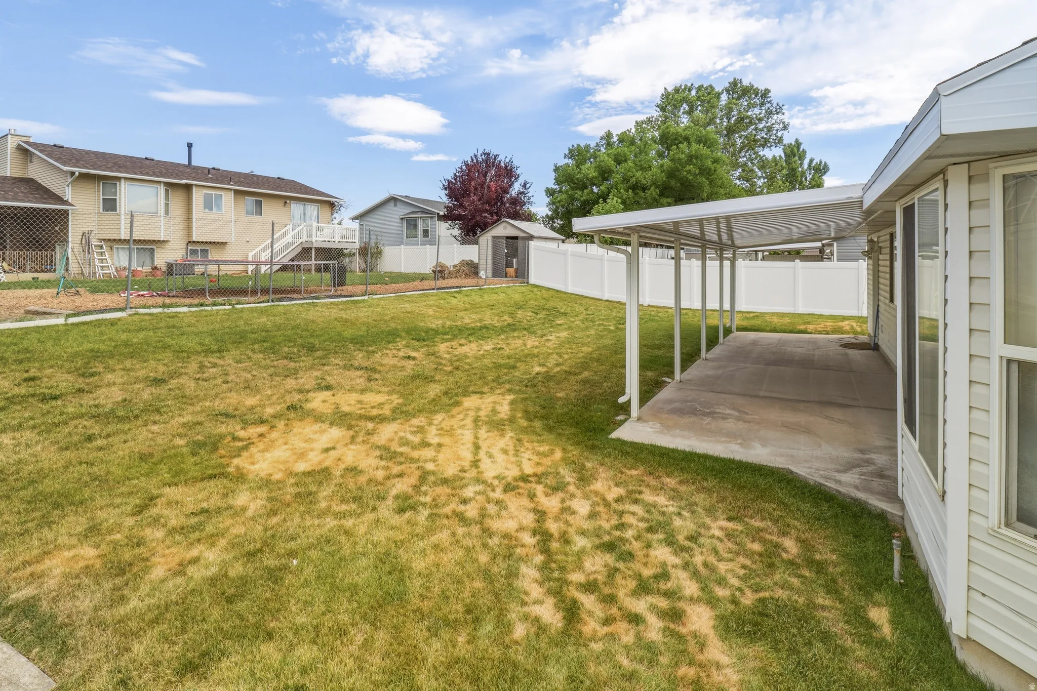 Expansive backyard featuring a covered concrete patio, white vinyl fencing, a detached storage shed, and a mature tree