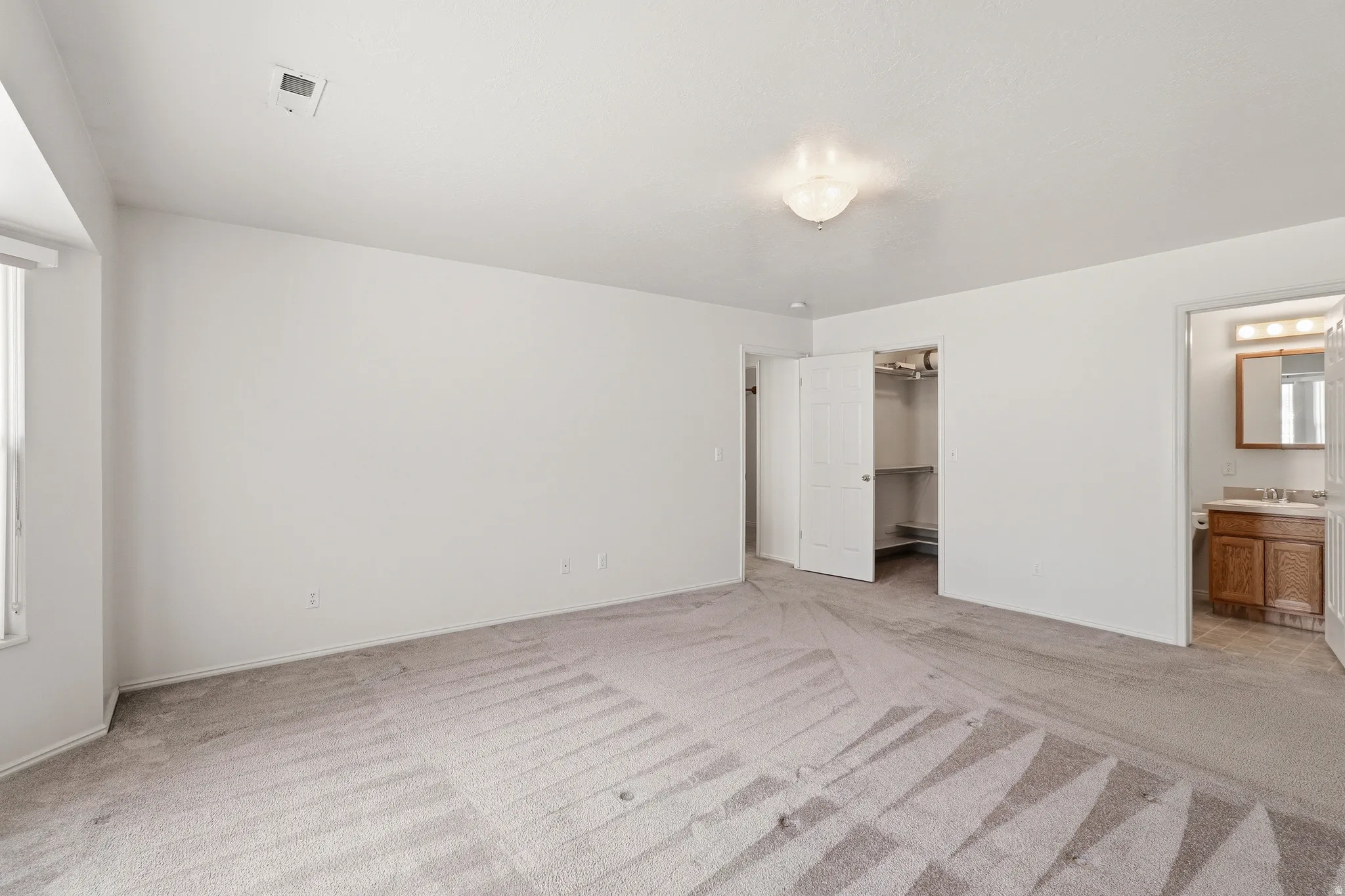 Spacious room with light gray carpeting, a window with vertical blinds, and a ceiling-mounted light fixture