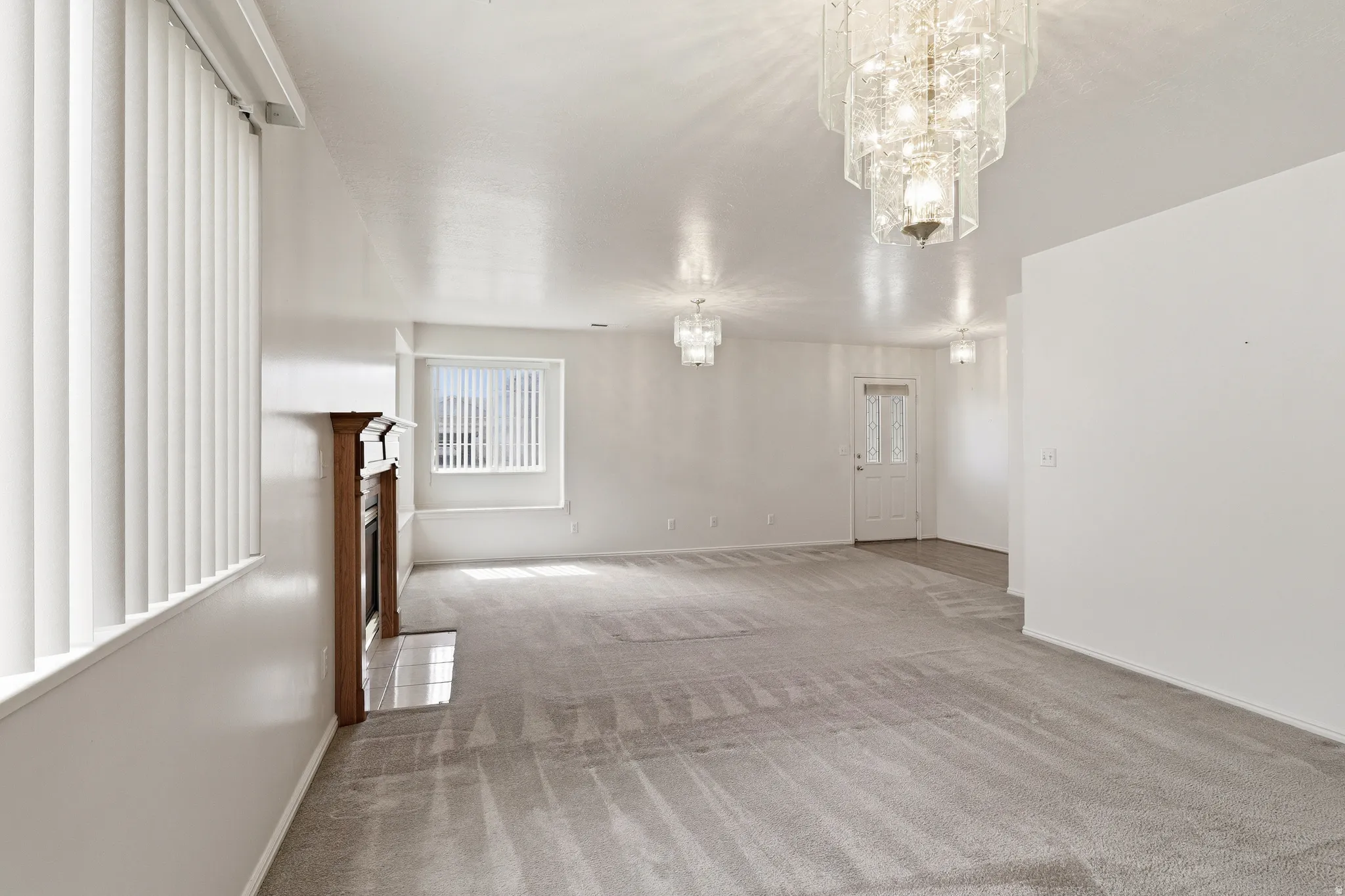 Spacious carpeted room featuring a decorative wood-finish fireplace, multiple vertical blinds, and a tiered crystal chandelier