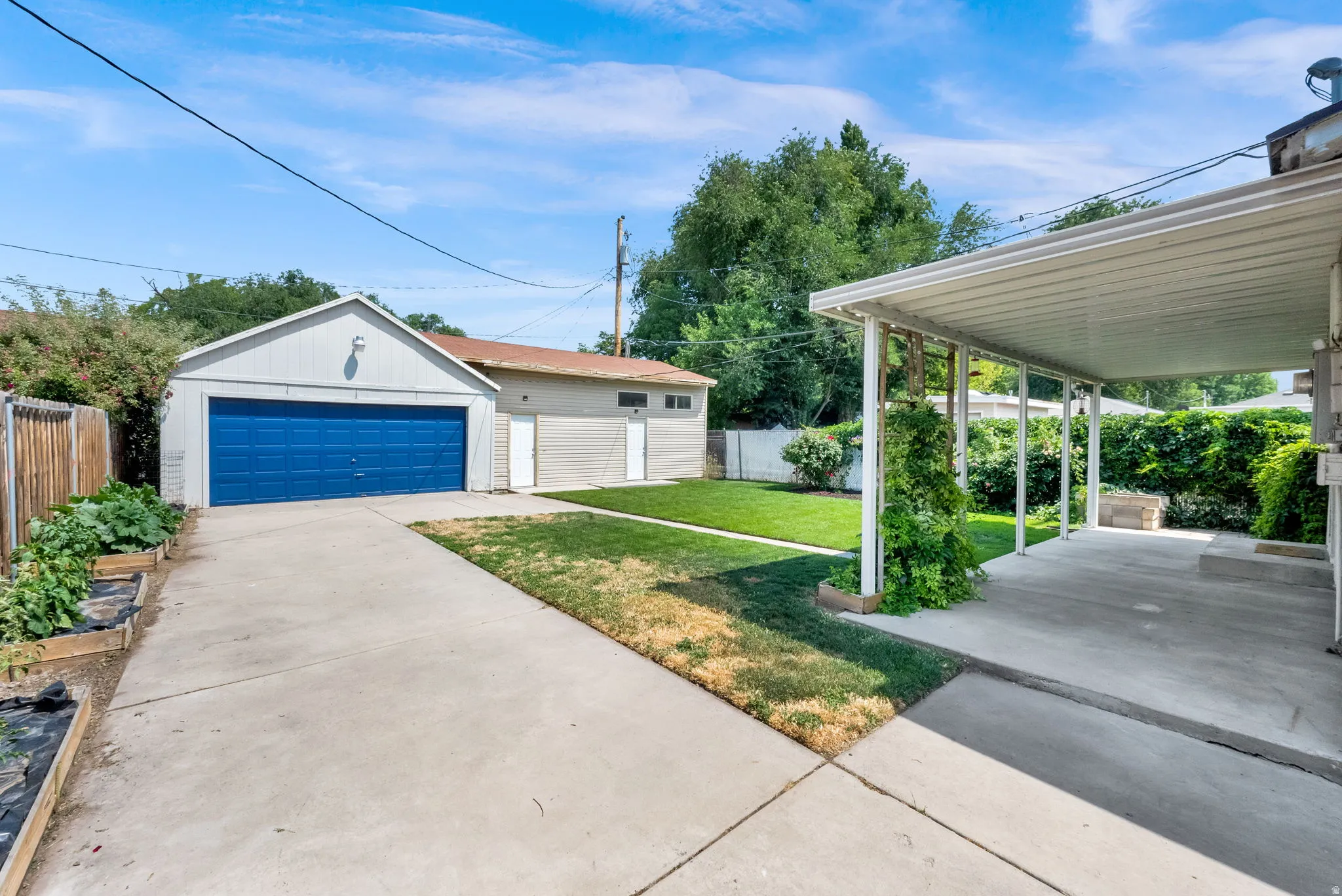 Detached garage with a vibrant blue door, ample concrete driveway, covered patio, manicured lawn, and raised garden beds