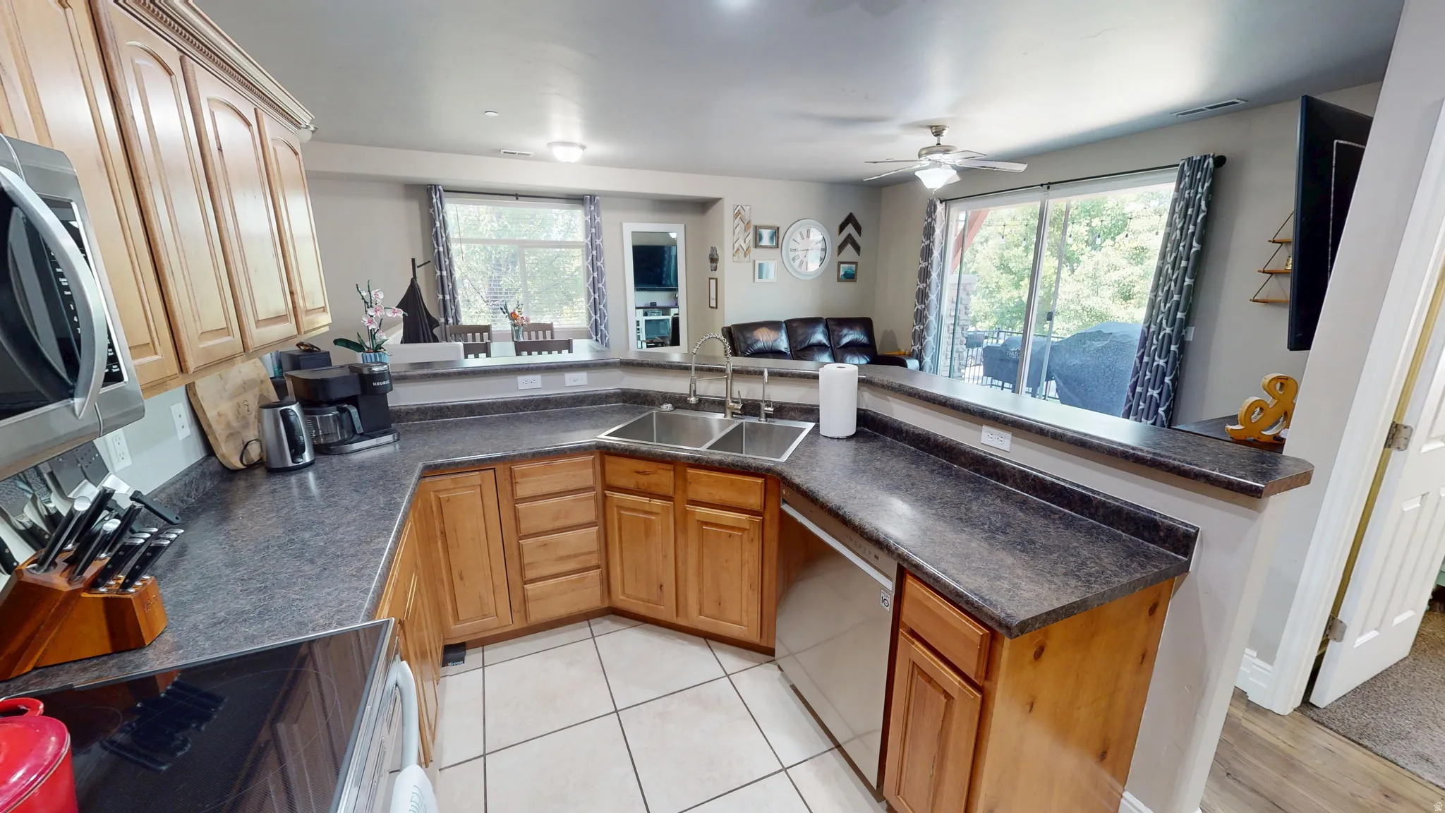 Open-concept kitchen featuring light wood cabinetry, dark countertops, a stainless steel double sink, and tile flooring