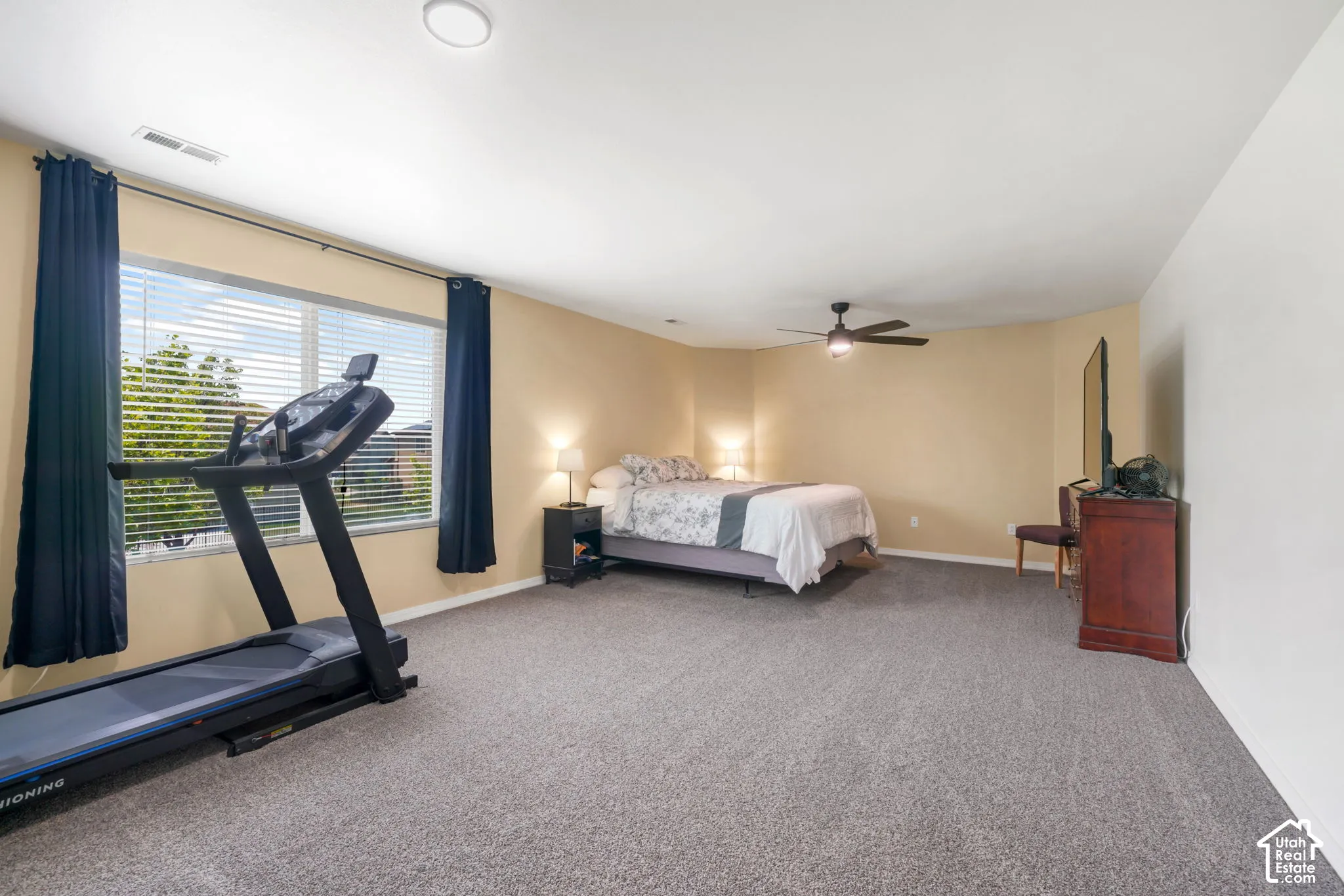 Spacious room featuring neutral-toned walls, gray carpeting, a large window with horizontal blinds, and a ceiling fan with integrated lighting
