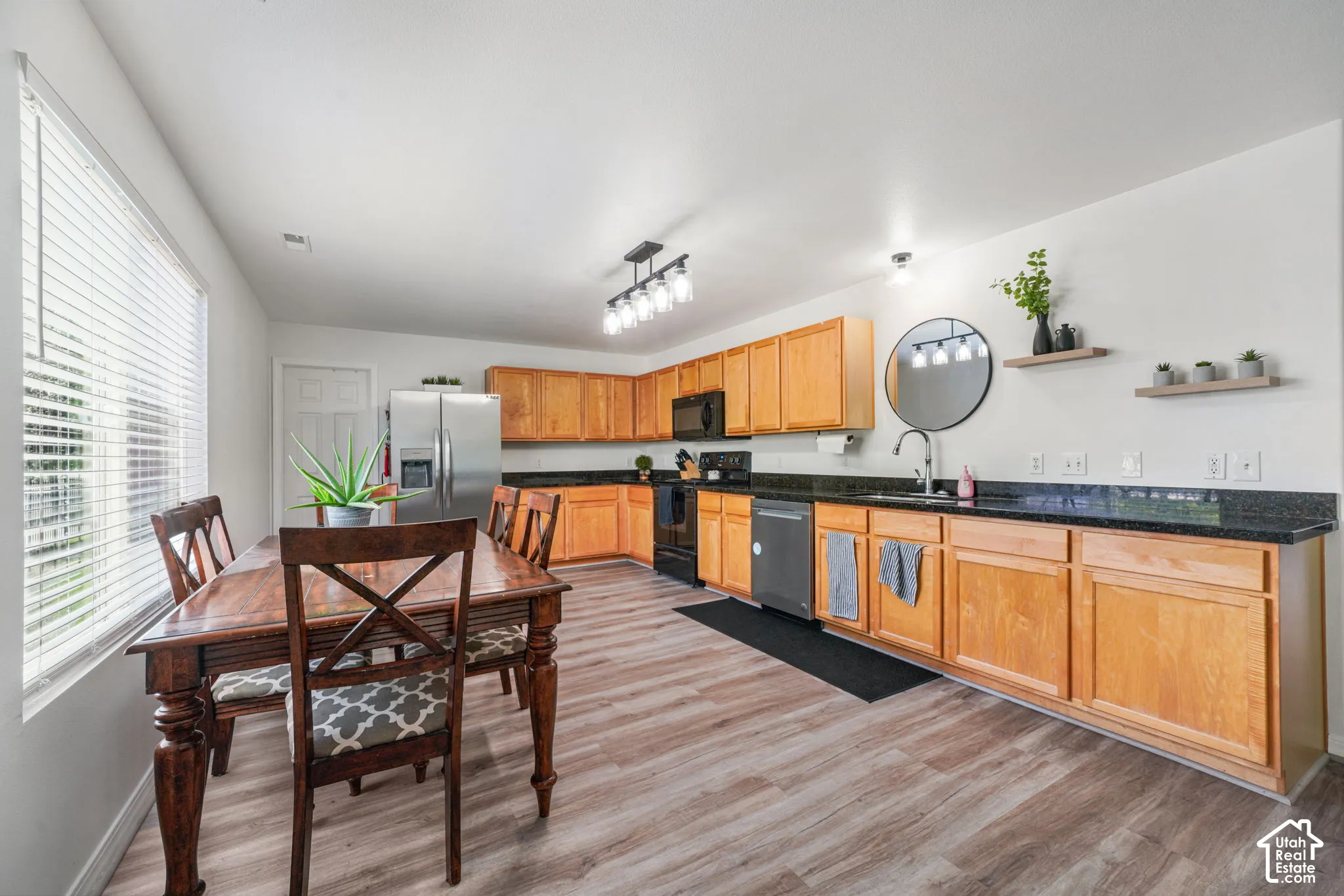 Open-concept kitchen and dining area featuring wood-finish flooring, light wood cabinetry, black countertops, and stainless steel appliances