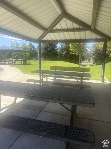 Covered picnic area featuring multiple metal picnic tables, a concrete slab foundation, and surrounding green space with mature trees
