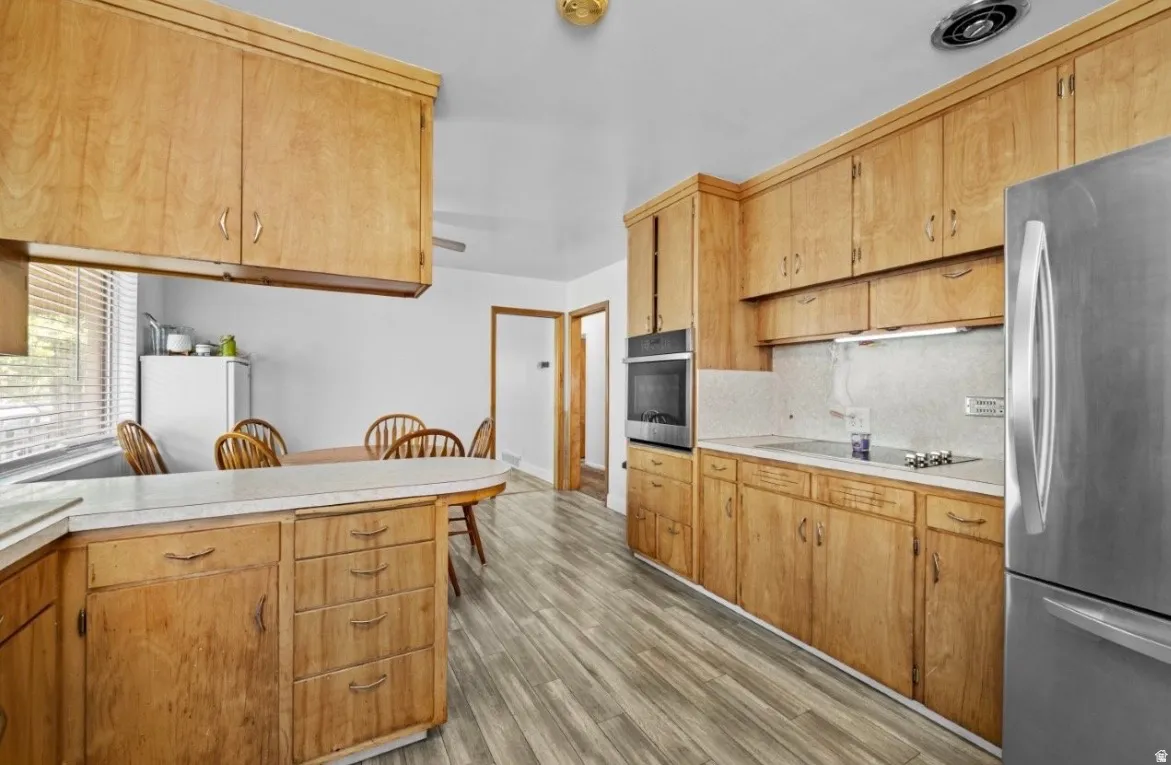Galley kitchen featuring wood cabinetry, light countertops, wood-finish flooring, stainless steel refrigerator, and a built-in oven