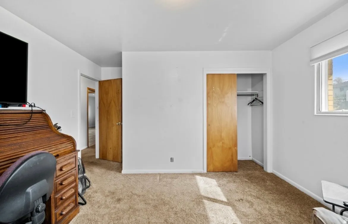 Carpeted room with white walls, featuring a window with a roller blind, and a wooden sliding closet door