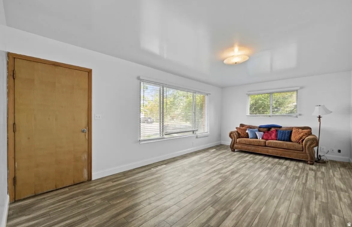 Spacious interior room featuring wood-finish flooring, white walls, and multiple large windows with blinds