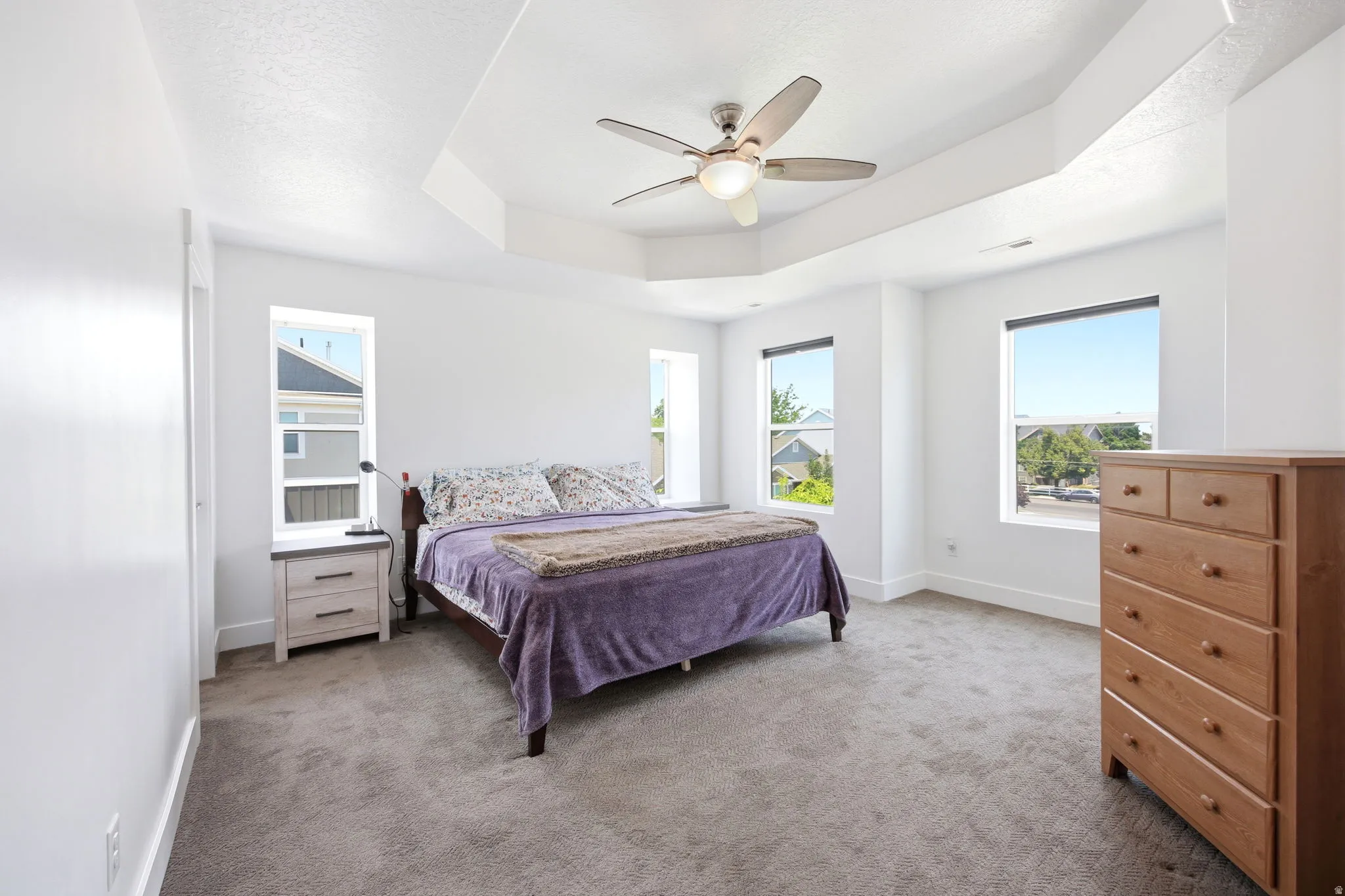 Spacious room featuring a tray ceiling with a flush-mount ceiling fan, four large windows, neutral carpeting, and white walls