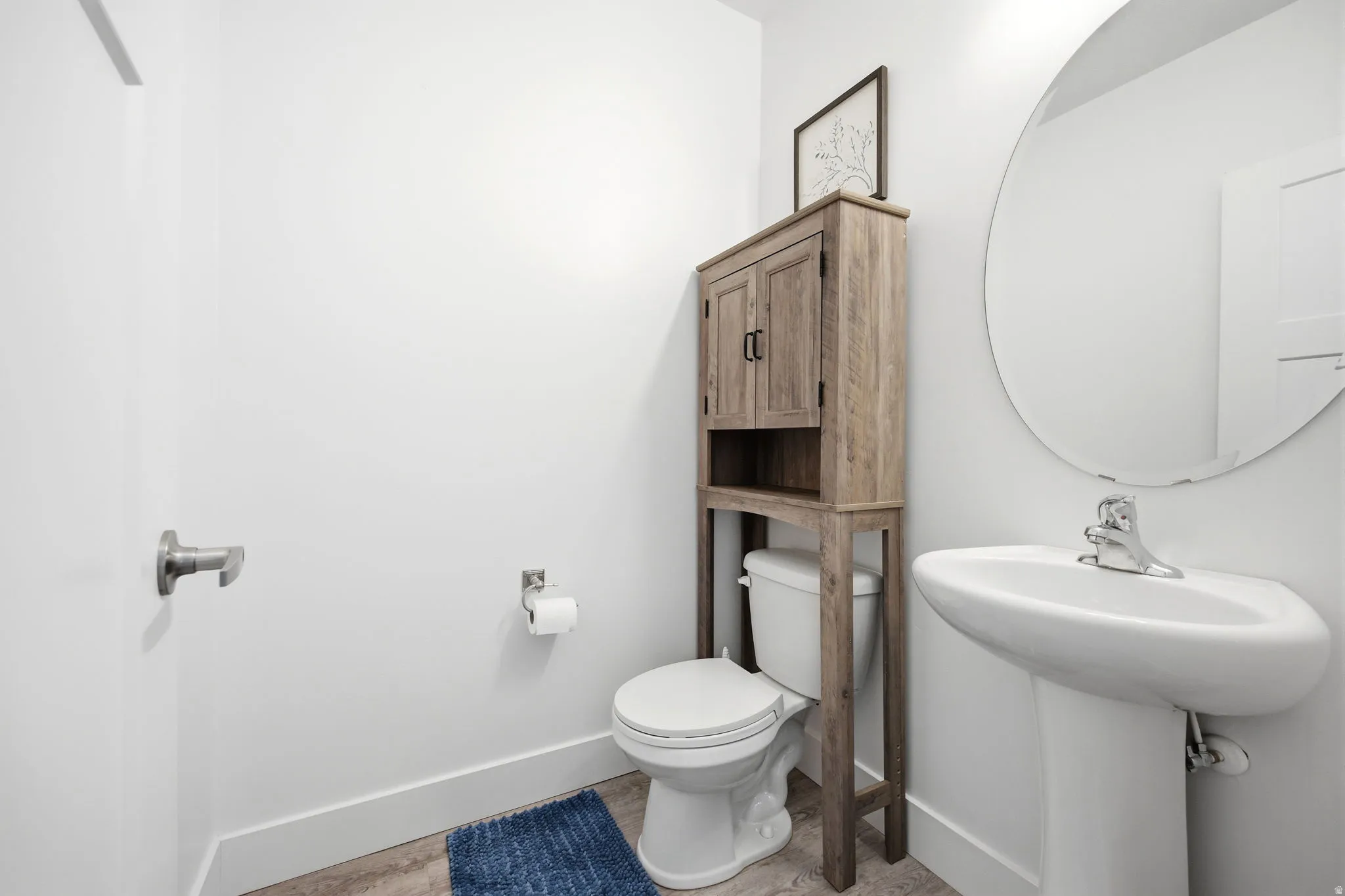 Pedestal sink with chrome faucet, circular wall mirror, and wood-finish over-the-toilet storage