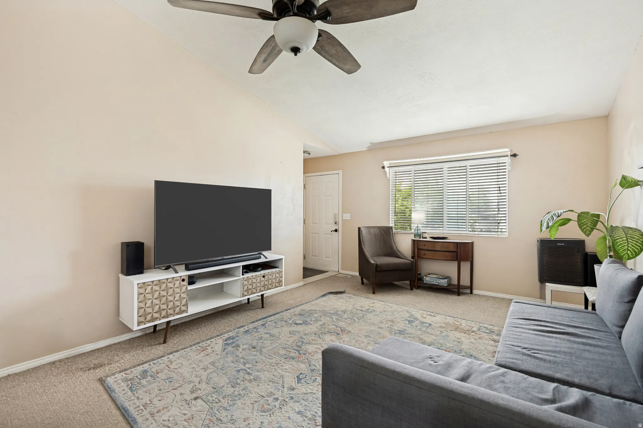 Living room featuring vaulted ceilings, a ceiling fan with integrated lighting, and a large window with horizontal blinds