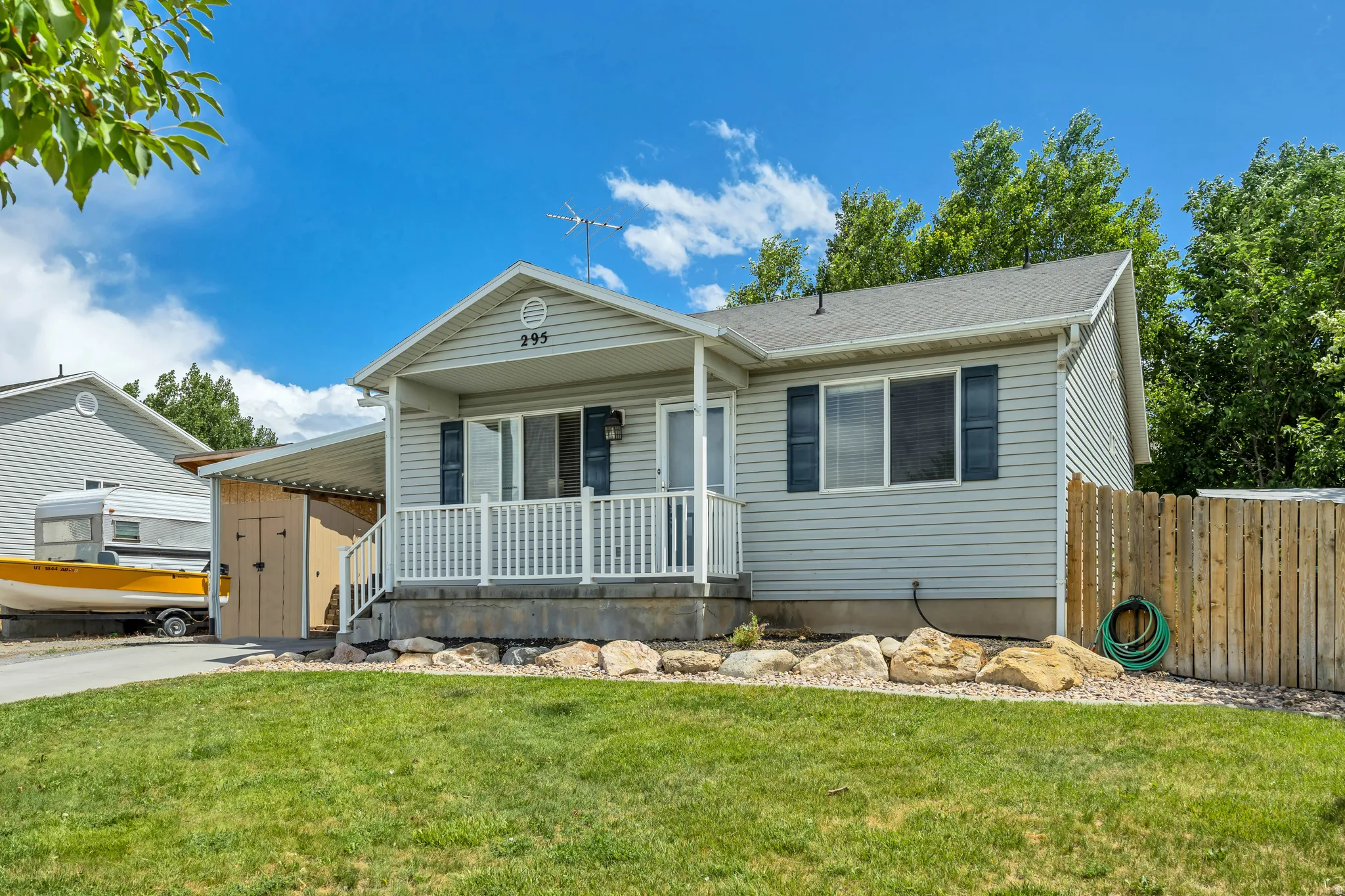 Light grey siding residence featuring a covered front porch with white railing, a wooden privacy fence, a paved driveway, and mature landscaping