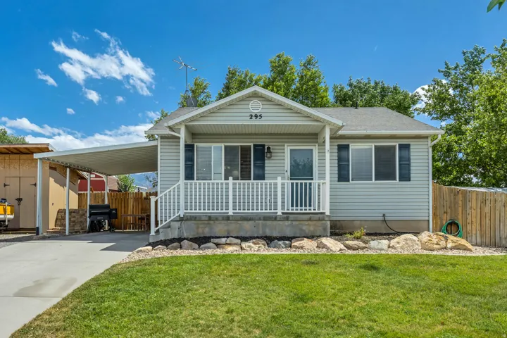 Gray siding exterior featuring a covered front porch with white railings, a paved driveway leading to a car canopy, a wooden fence, and a front lawn with rock landscaping