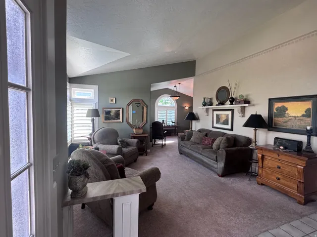 Spacious living area featuring vaulted ceilings, plantation shutters on multiple windows, neutral carpeting, and a built-in wall shelf