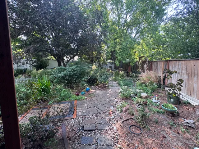 Expansive garden area featuring a stone paver pathway, diverse plantings, mature trees, and a wood privacy fence