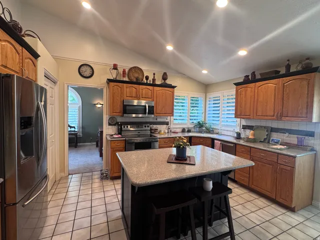 Kitchen featuring a central island with a granite-style countertop, stainless steel appliances, wood-finish cabinetry, tile flooring, and plantation shutters