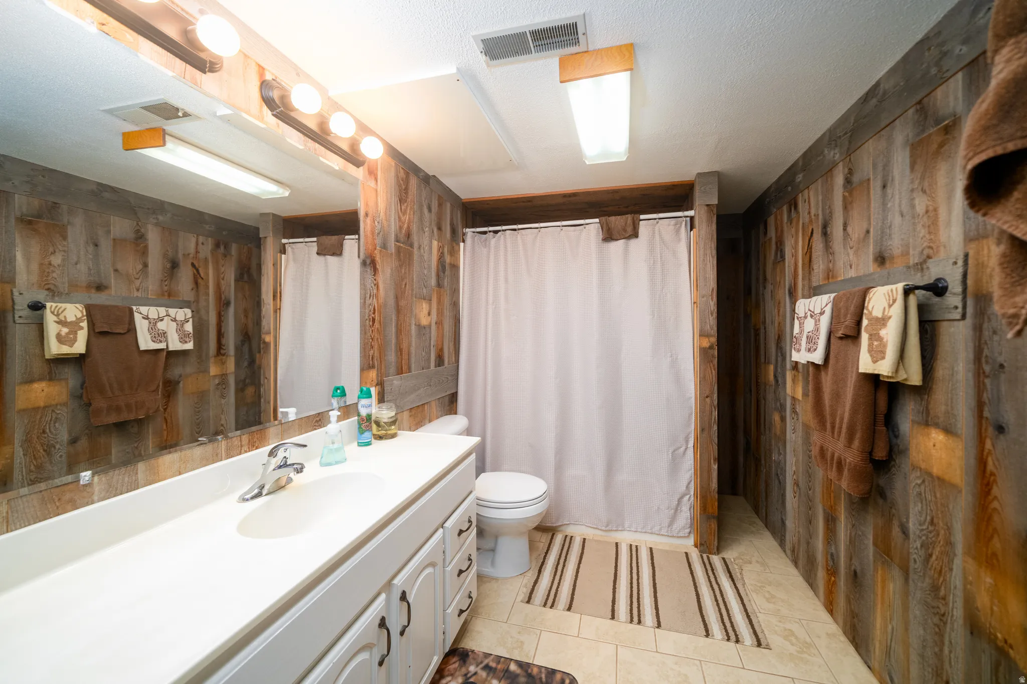 Bathroom featuring wood-finish plank walls, a single vanity with a white countertop, a mirror with strip lighting, a toilet, and a tiled shower area