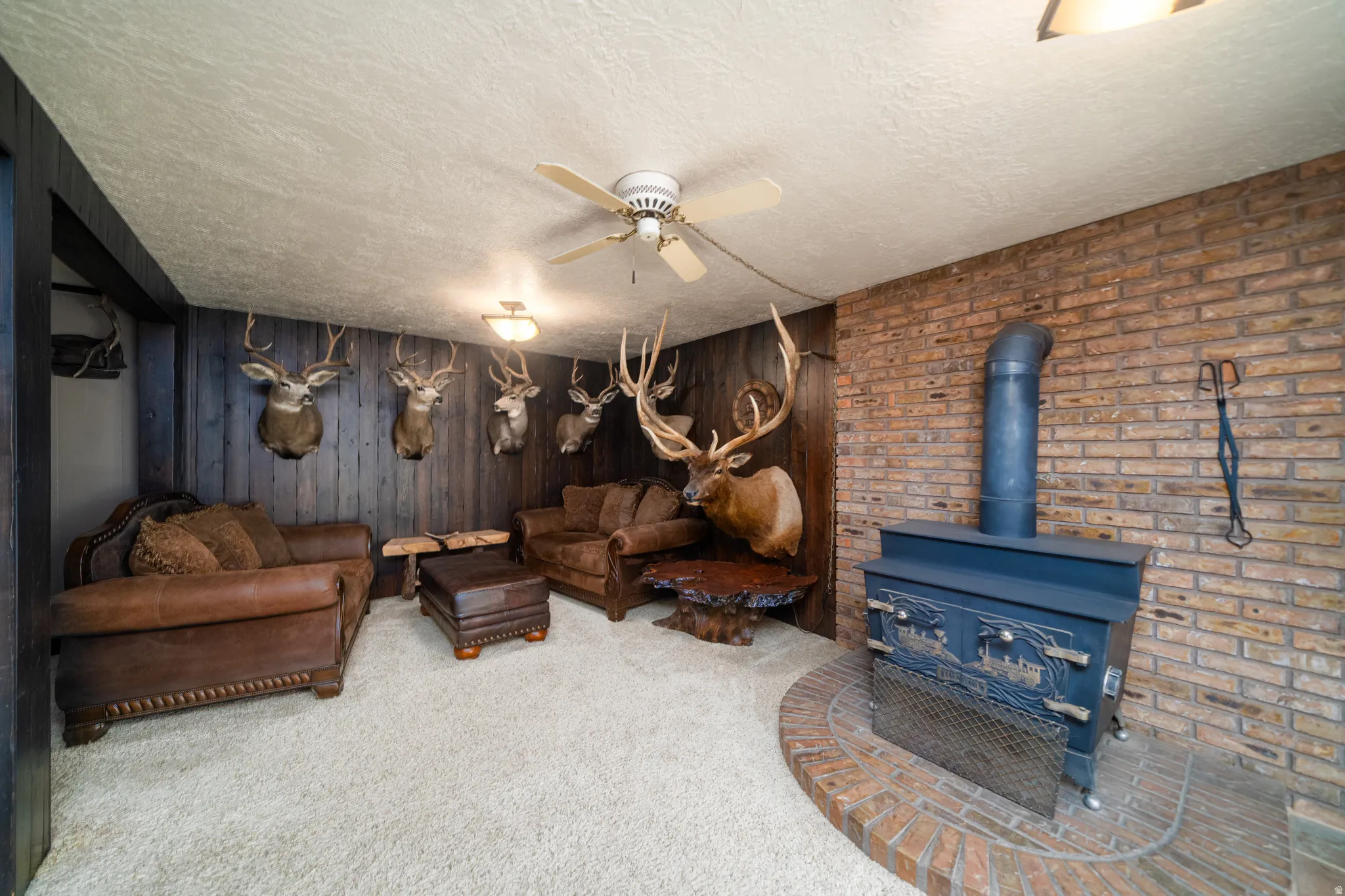 Carpeted recreation room featuring an exposed brick wall, wood-paneled wall, and a corner wood-burning stove with a brick hearth
