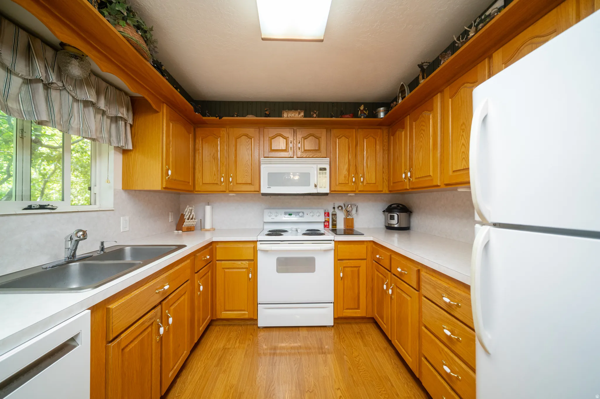 U-shaped kitchen featuring wood cabinetry, white countertops, a double basin stainless steel sink, a window with outdoor tree scenery, and white appliances including a refrigerator, range, and microwave