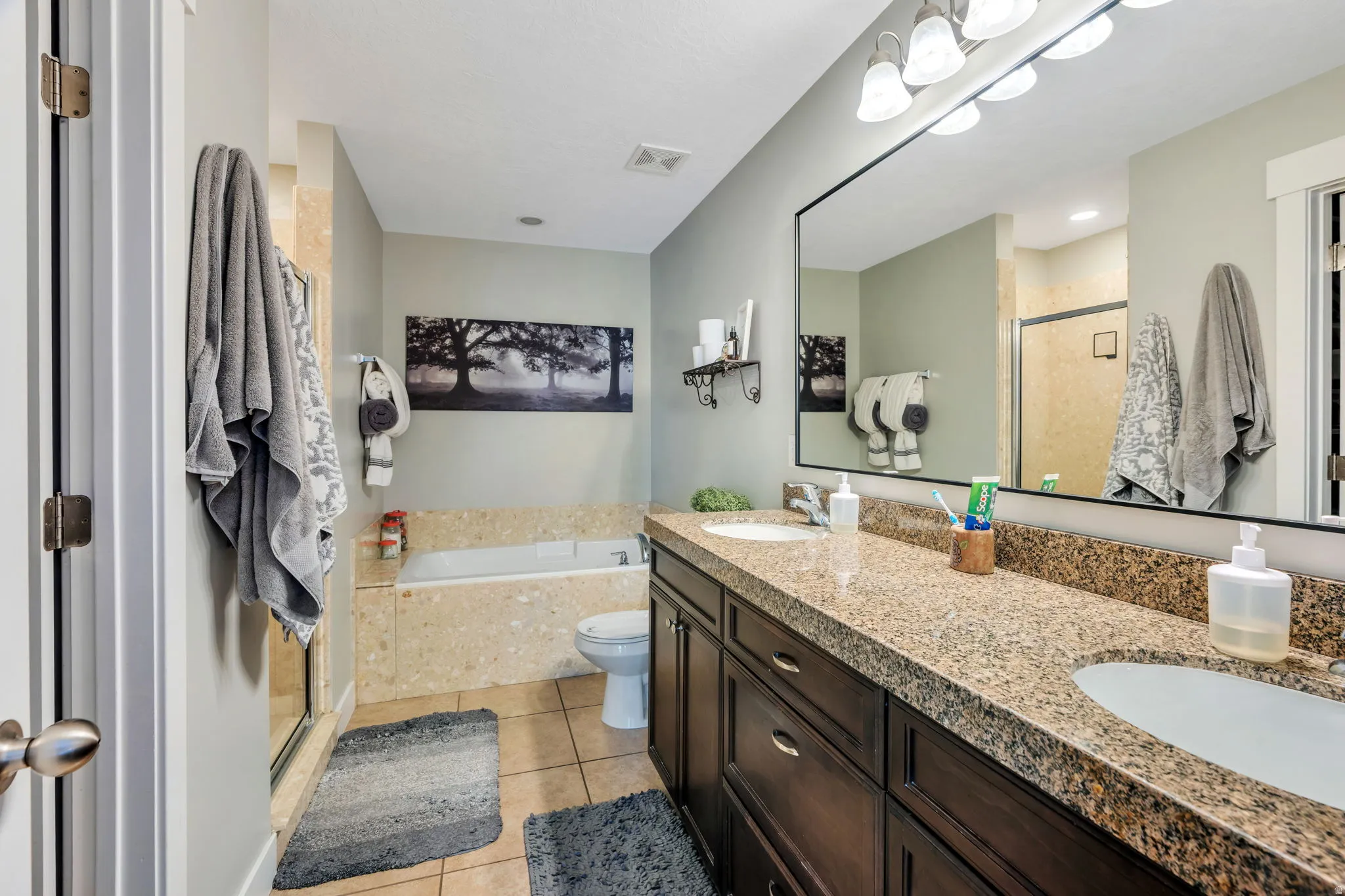 Primary Bathroom featuring a dual-sink vanity with granite-style countertops, dark wood cabinetry, a large frameless mirror, and overhead sconce lighting