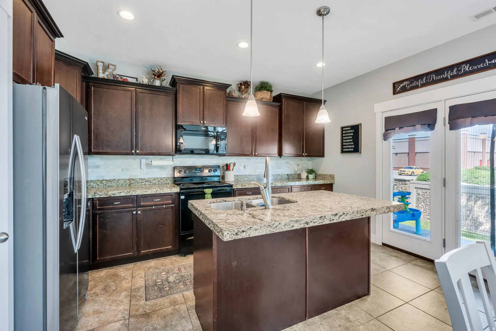 Kitchen featuring dark wood cabinetry, a central island with a sink, stainless steel refrigerator, black range, and double glass-paneled doors