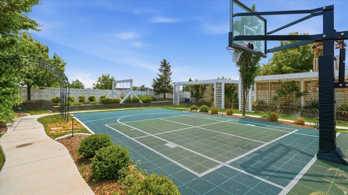 Outdoor sports court with a basketball hoop, surrounded by landscaped garden beds, a winding concrete pathway, a decorative metal archway, and white lattice privacy fencing