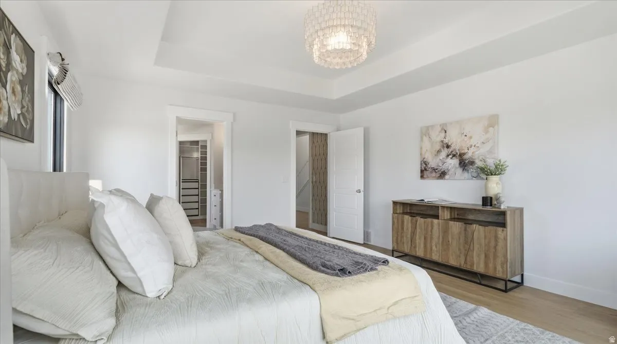 Spacious bedroom featuring a tray ceiling with a chandelier, wood-finish flooring, and bright white walls