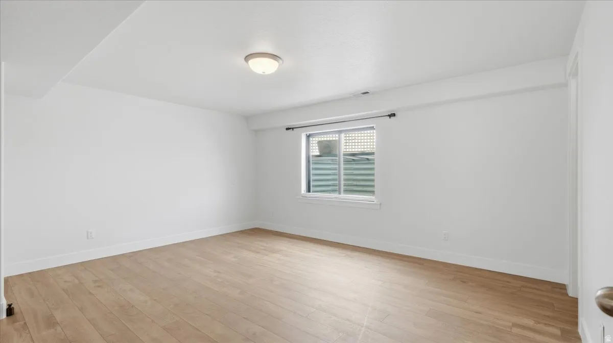 Spacious room featuring light wood-finish flooring, white walls, a flush-mount ceiling light fixture, and a window with a window well