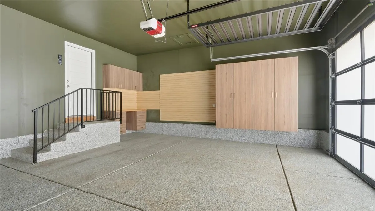 Spacious garage featuring a finished epoxy floor, built-in wood-finish cabinetry, slat wall storage system, an overhead storage rack, and a glass-paneled garage door