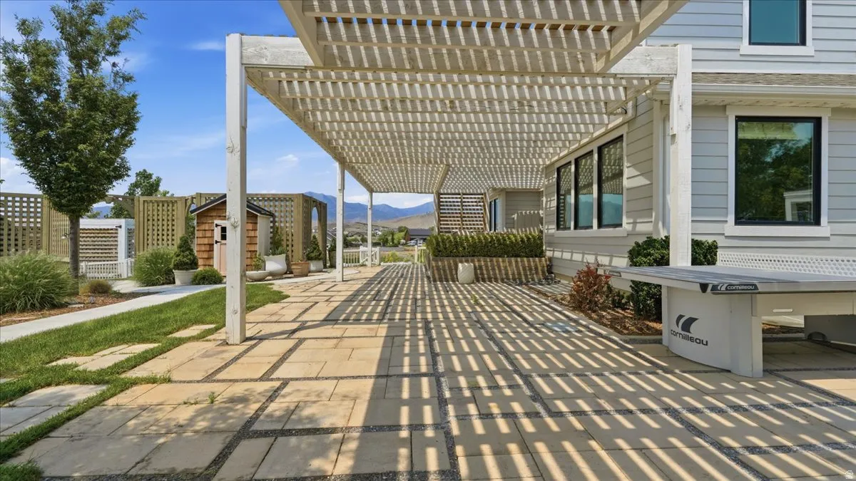 Expansive outdoor patio featuring a slatted pergola, stone paver flooring, manicured lawn, and a wooden storage shed