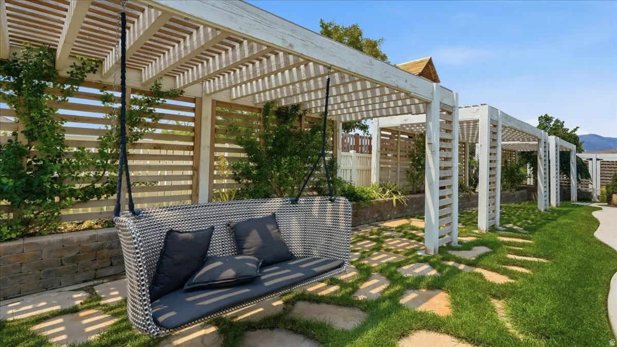 Outdoor pergola walkway featuring a suspended swing bench, stone paver pathway, manicured lawn, and slatted privacy fencing
