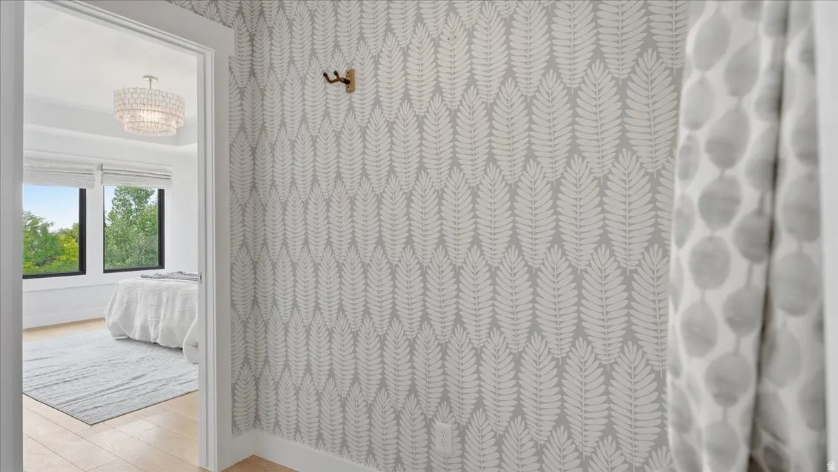 Light wood-finish flooring extends from the hallway into the room, featuring a tiered crystal chandelier, expansive windows, and a decorative botanical wallpaper accent wall