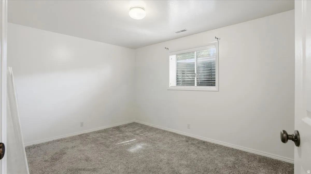 Spacious room featuring neutral carpet flooring, white walls, a single window with horizontal blinds, and a ceiling-mounted light fixture