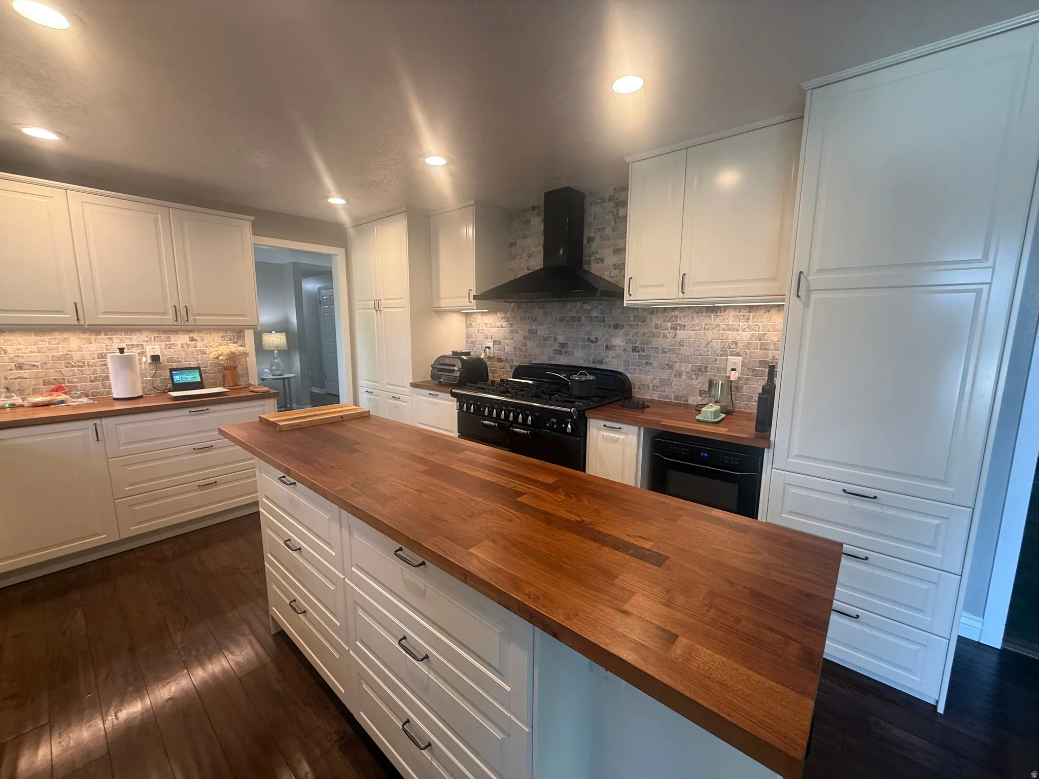 Central kitchen island with wood-finish countertop, dark wood-finish flooring, white cabinetry, light-toned brick backsplash, and black range hood