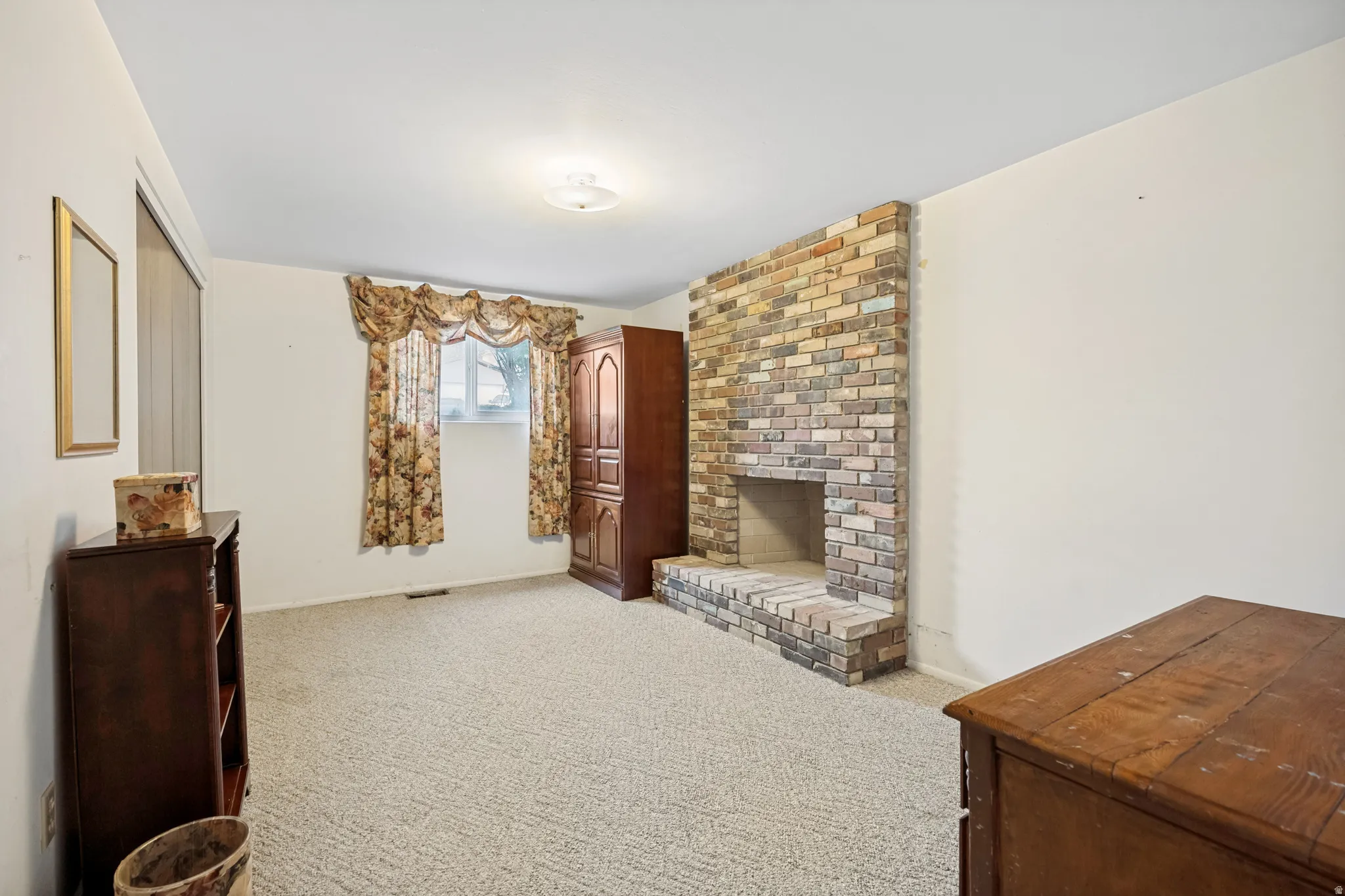 Spacious room featuring a brick fireplace with a raised hearth, light-colored carpet, and a window providing natural light