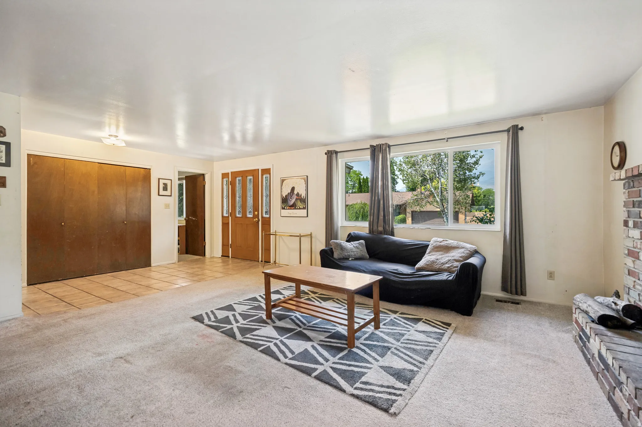 Spacious living area featuring a large picture window, brick fireplace with a hearth, and warm wood-finish double entry doors
